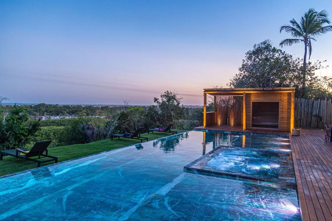 Piscina de borda infinita e jacuzzi ao entardecer, com a sauna iluminada